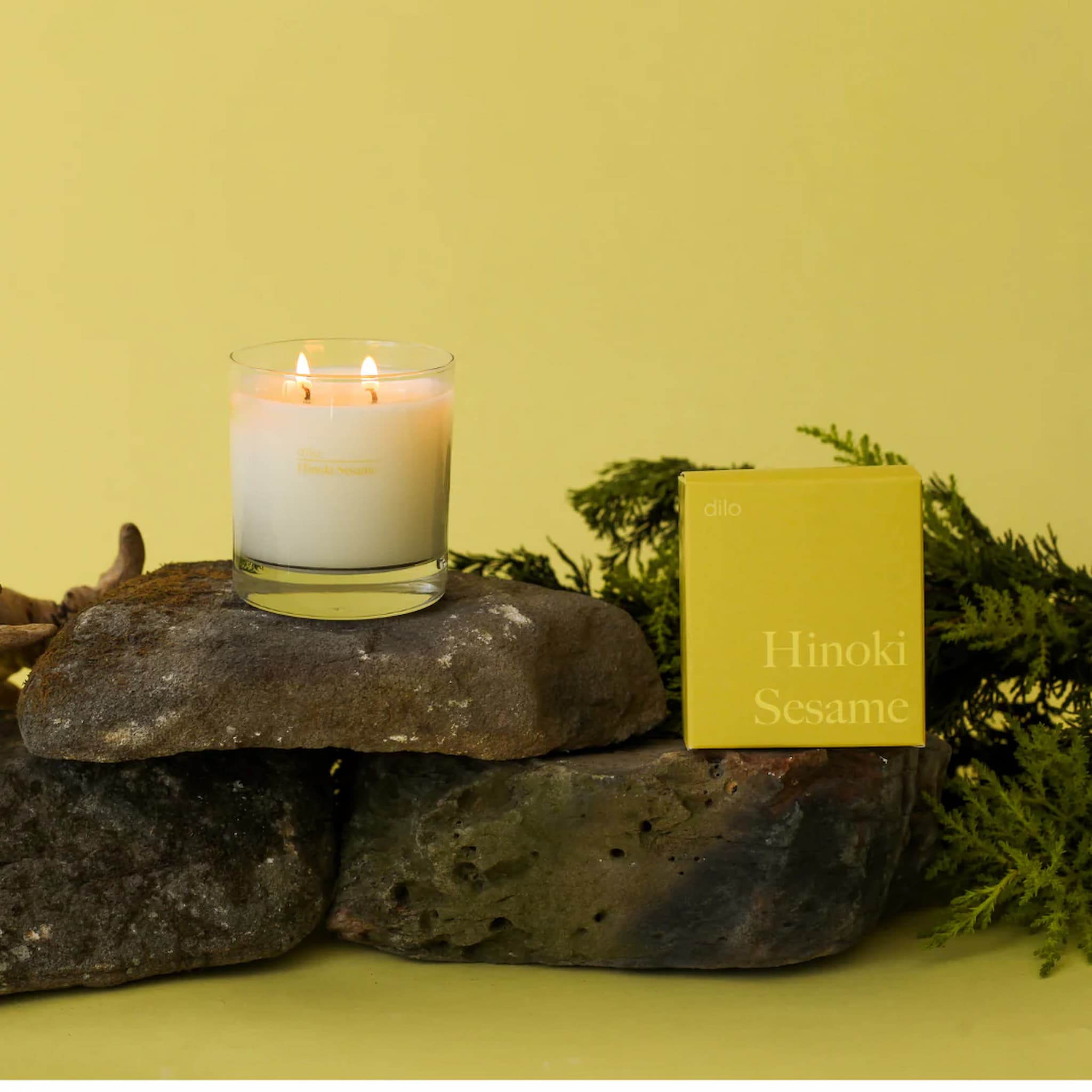 Dilo candle in a glass holder on a rock with a yellow box labeled 'Hinoki Sesame' against a yellow background