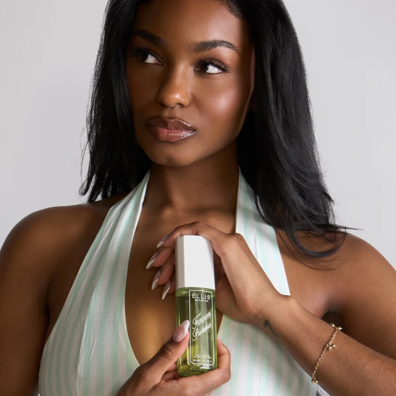 Woman holding an Ellis Brooklyn Forever Pistachio perfume mist bottle against a neutral background.