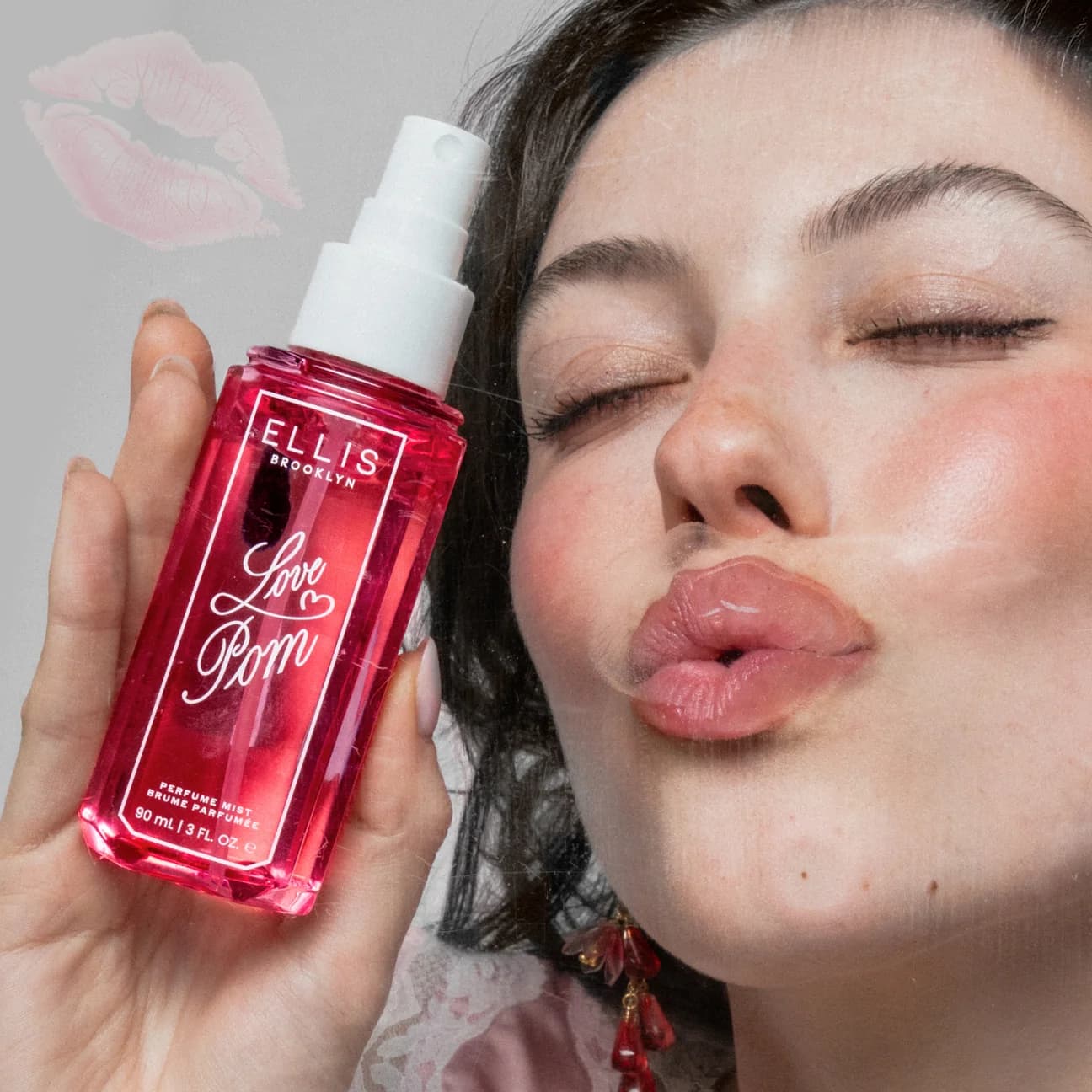 A close-up lifestyle shot of a woman holding a bottle of Ellis Brooklyn Love Pom perfume mist. The woman is puckering her lips, and a pink lip print graphic is visible in the top left corner.