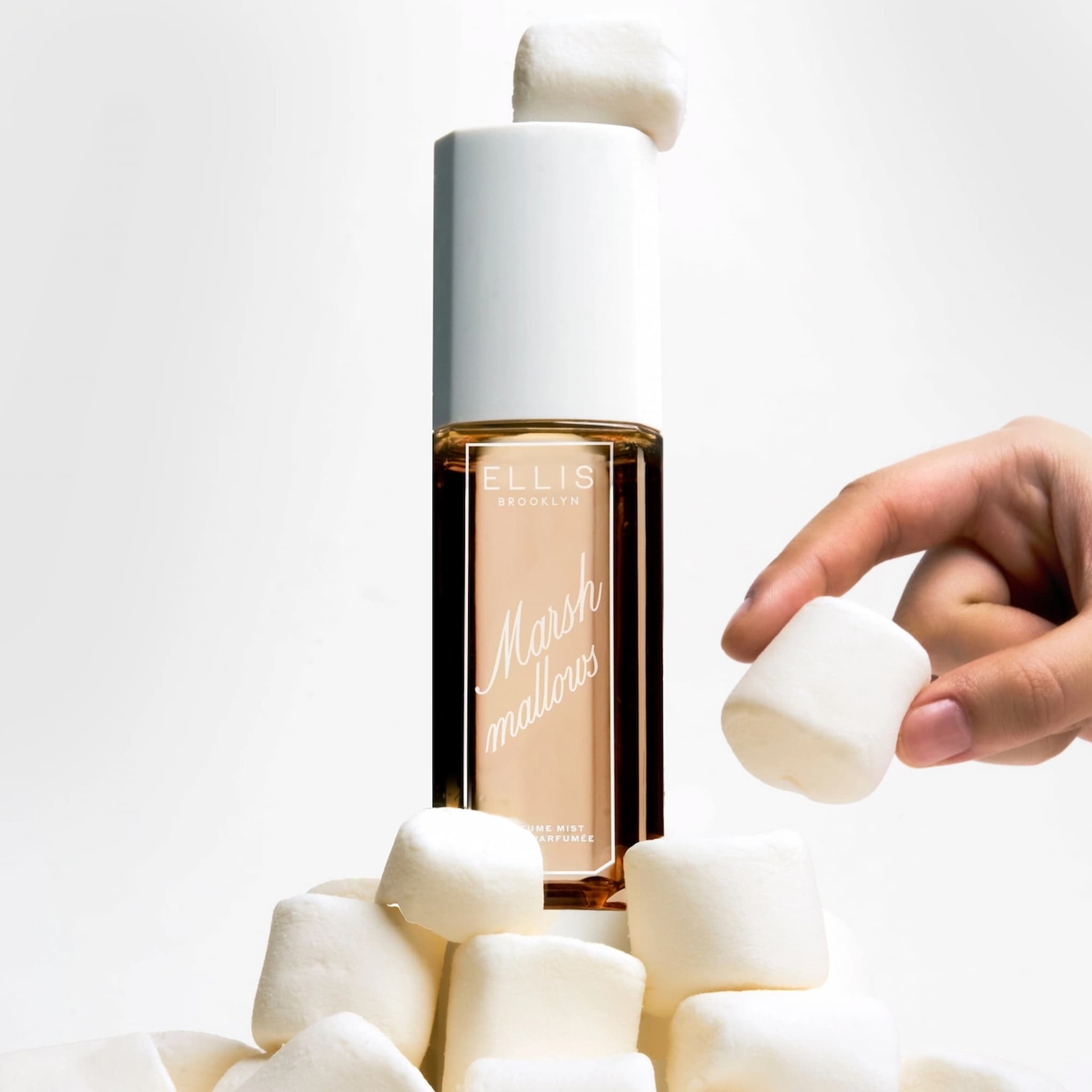 Ellis Brooklyn Marshmallows Perfume Mist sitting on a tower of marshmallows next to a hand holding a marshmallow.