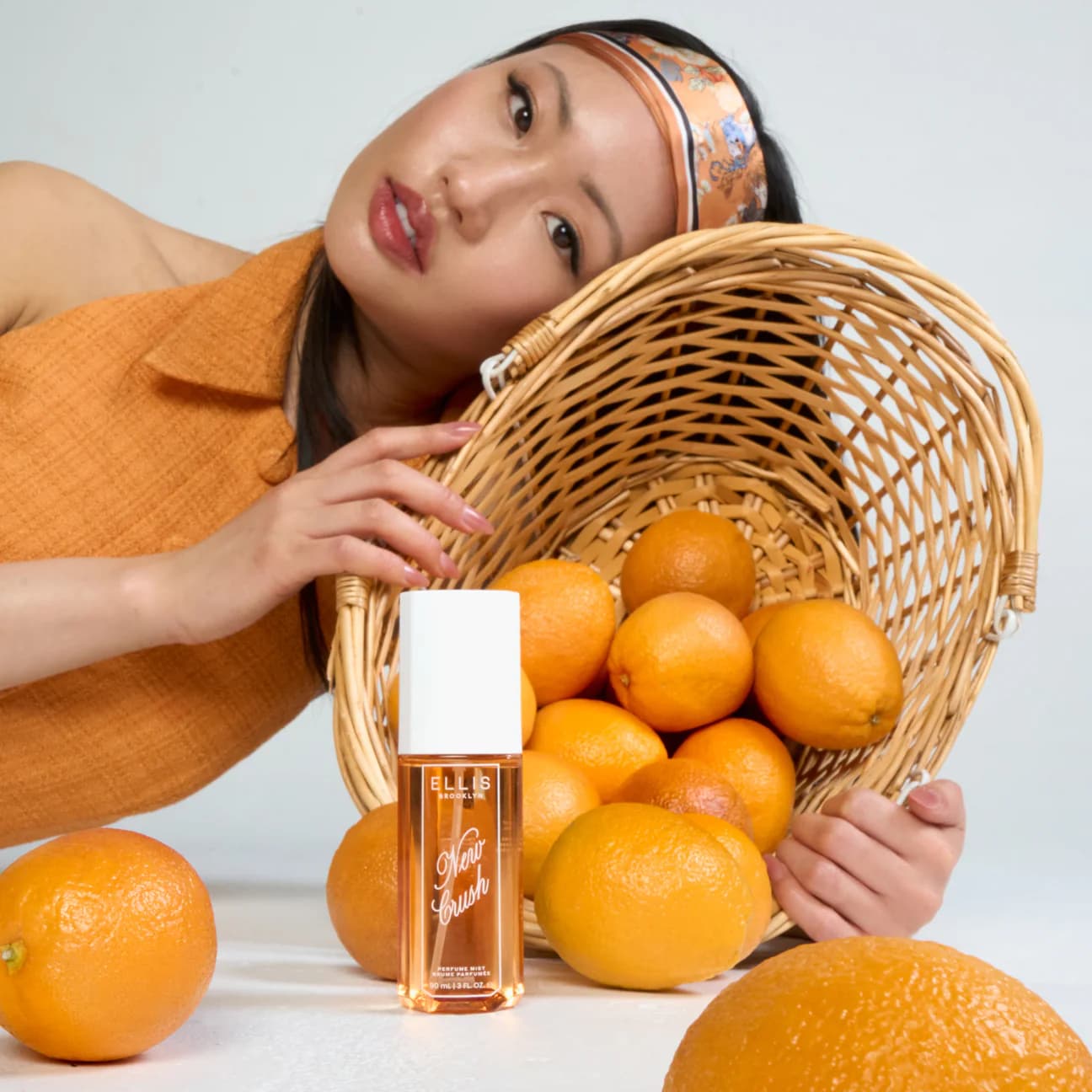 Woman with a basket of oranges and a bottle of Ellis Brooklyn New Crush perfume mist on a light background.
