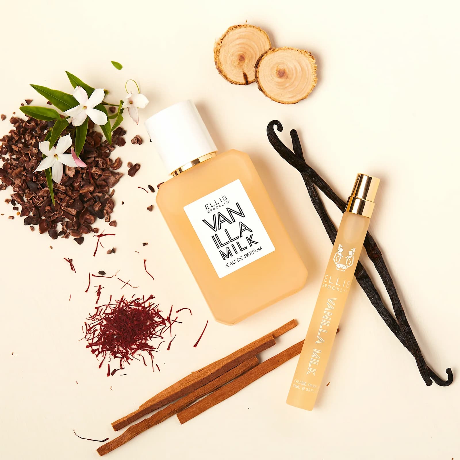 Two different size bottles of Ellis Brooklyn Vanilla Milk against a cream-colored background, surrounded by various ingredients found in the perfume, including vanilla bean, cocoa, wood, and flowers.