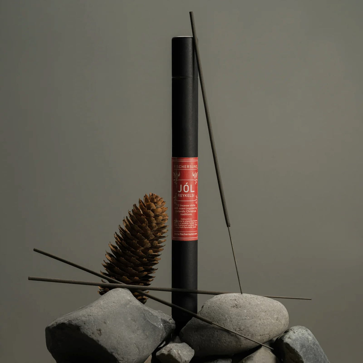 A black cylindrical incense stick holder with a red label reading Fischersund JÖL incense stands upright on stacked gray stones, with incense sticks and a pinecone arranged around it against a plain taupe background.