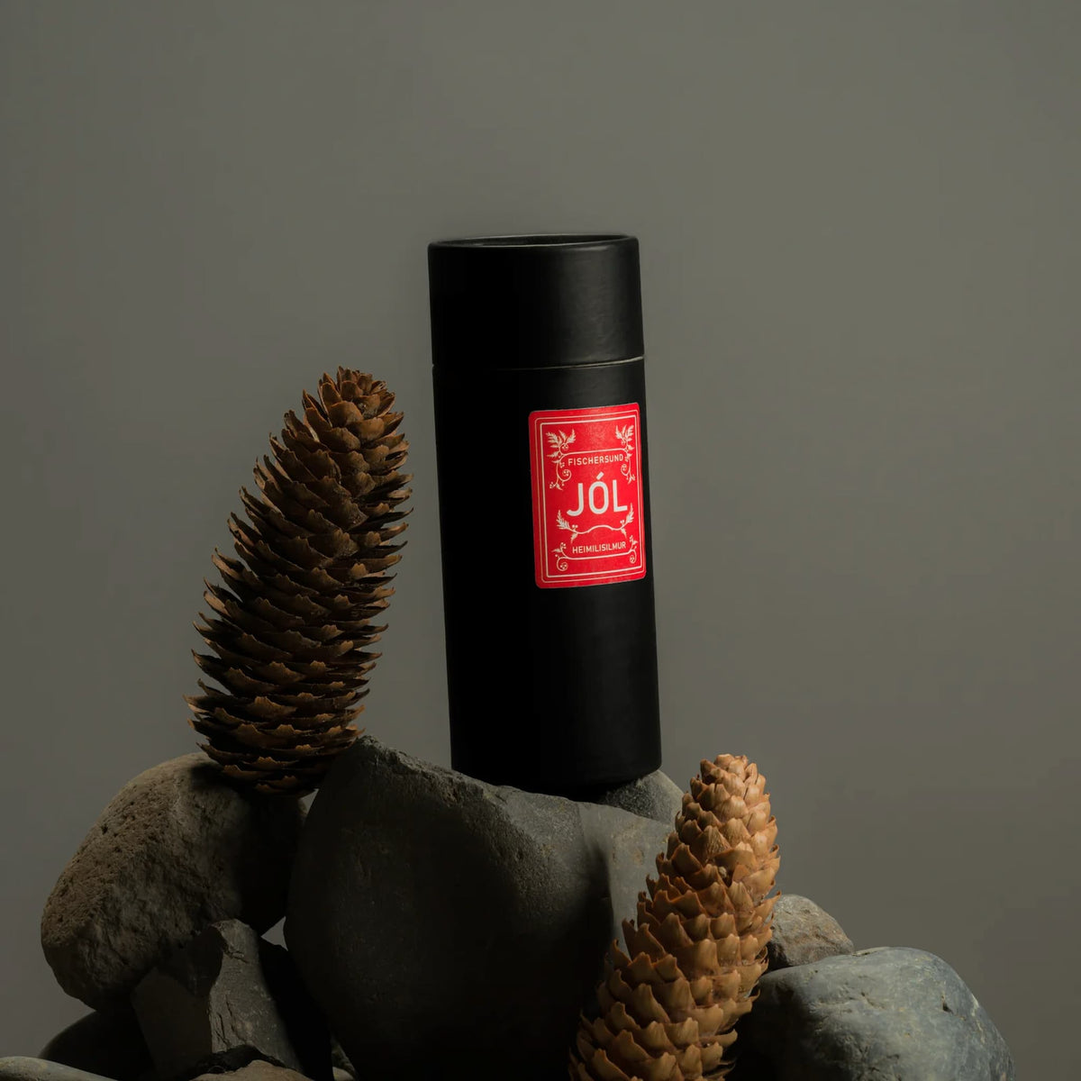 Black cylindrical container for Fischersund Jol room spray with a red label on a rock with pine cones against a gray background