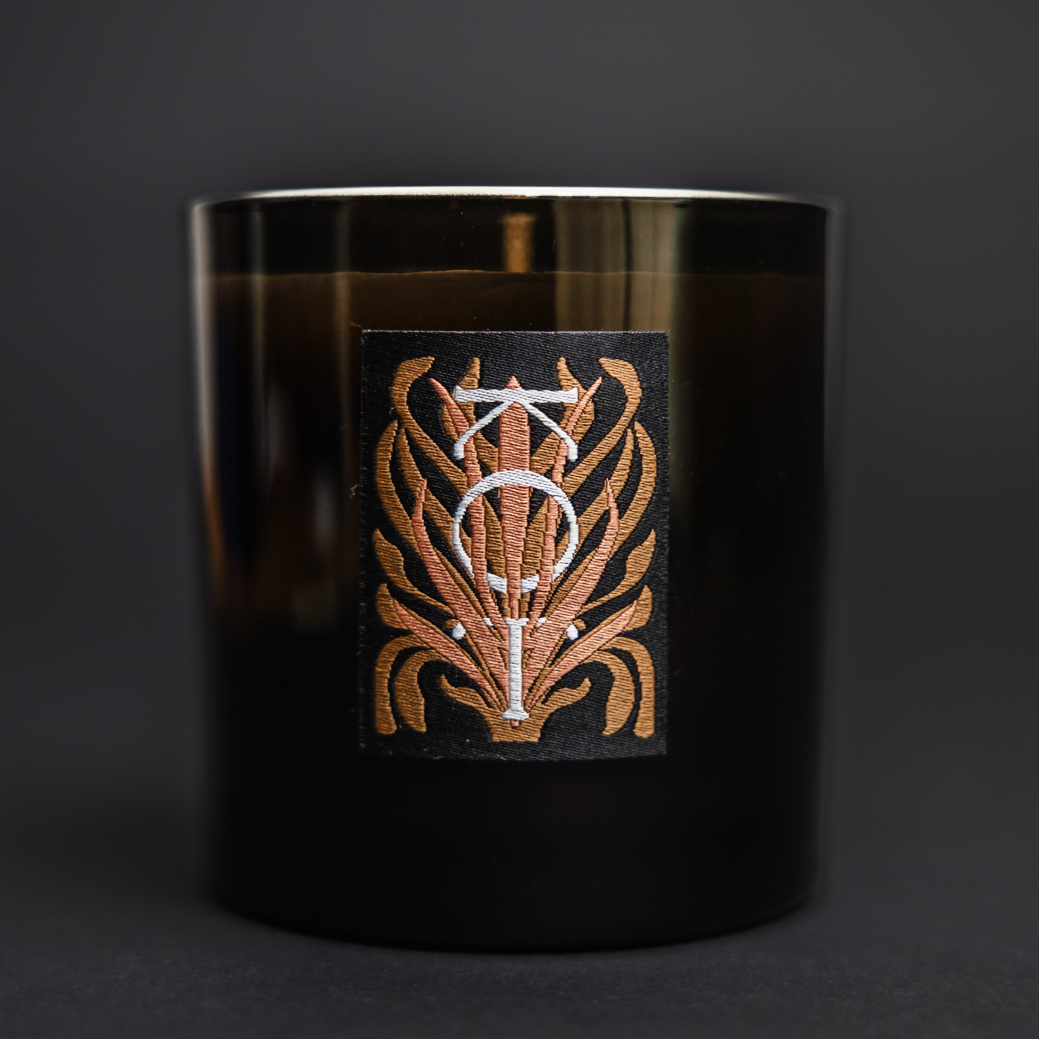 The Fischersund KOT black glass jar candle with a metallic gold and white decorative label featuring a stylized monogram, set against a dark gray background.