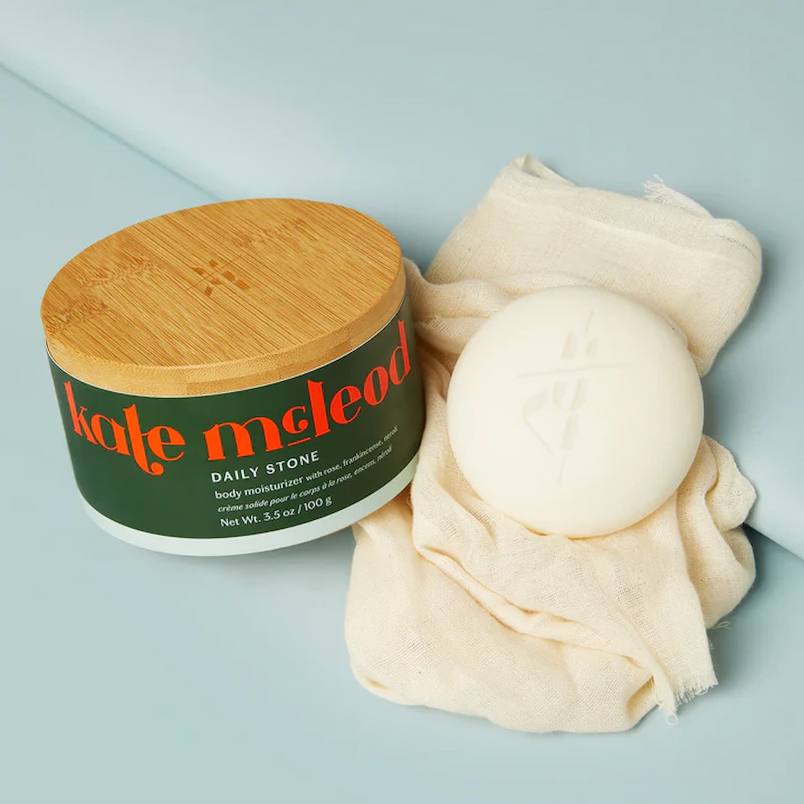 A white bar of Kate McLeod Daily Stone sits on top of a cream-colored fabric, next to the wooden box for the product.