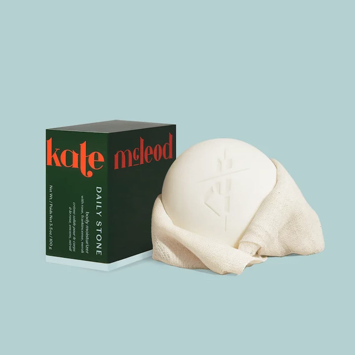 A white bar of Kate McLeod Daily Stone sits wrapped in a cream-colored fabric next to the green packaging for the product.