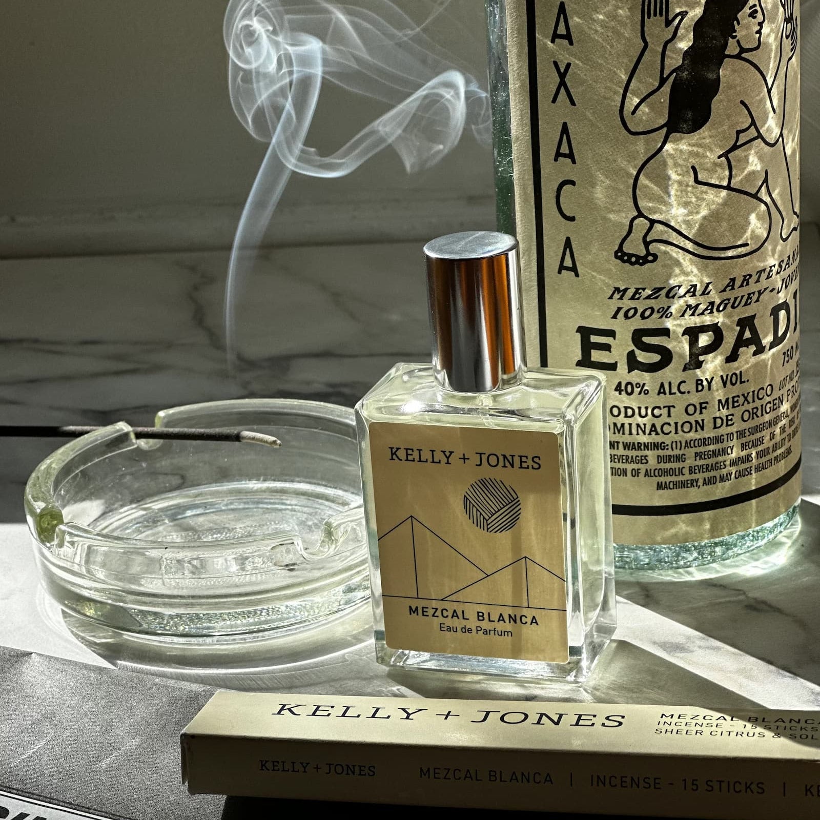 Kelly + Jones Mezcal Blanca Eau de Parfum perfume bottle displayed on a marble surface next to a smoking incense stick, and a large bottle of Oaxaca Espadilte Mezcal.