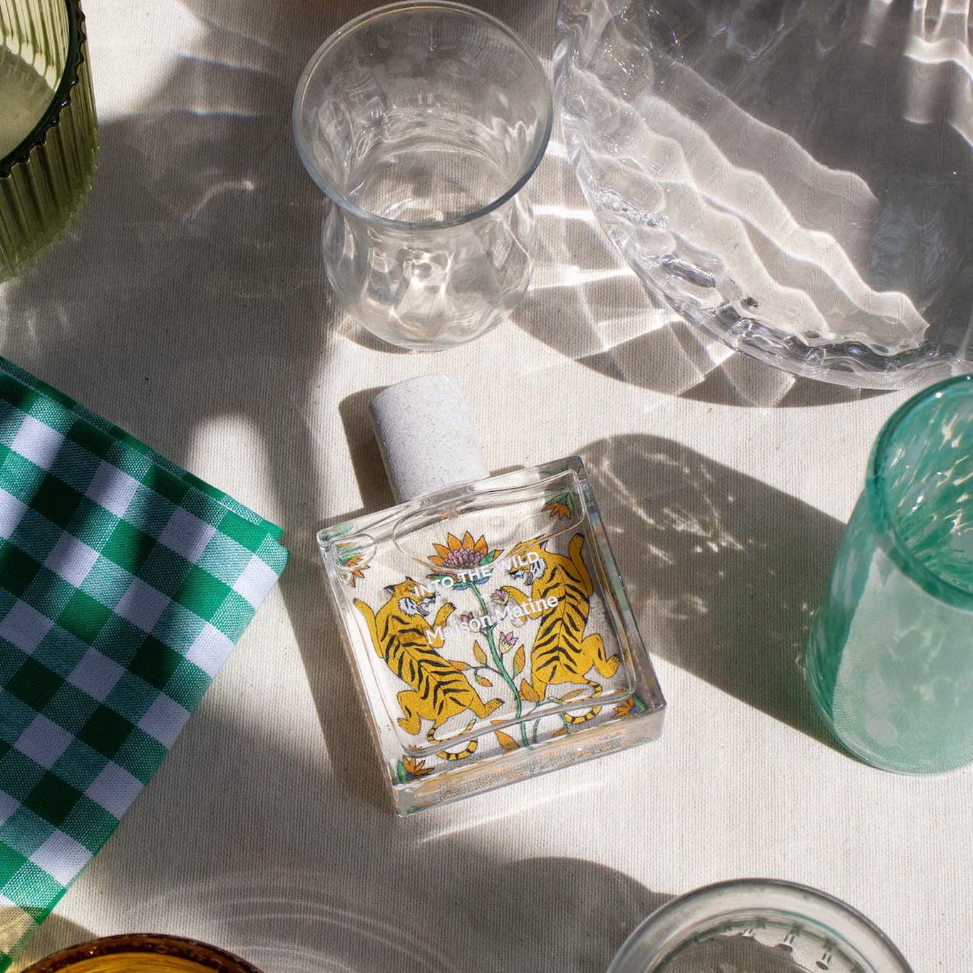 Maison Matine Into the Wild perfume bottle on a cream colored table with a turquoise glass and gingham napkin.