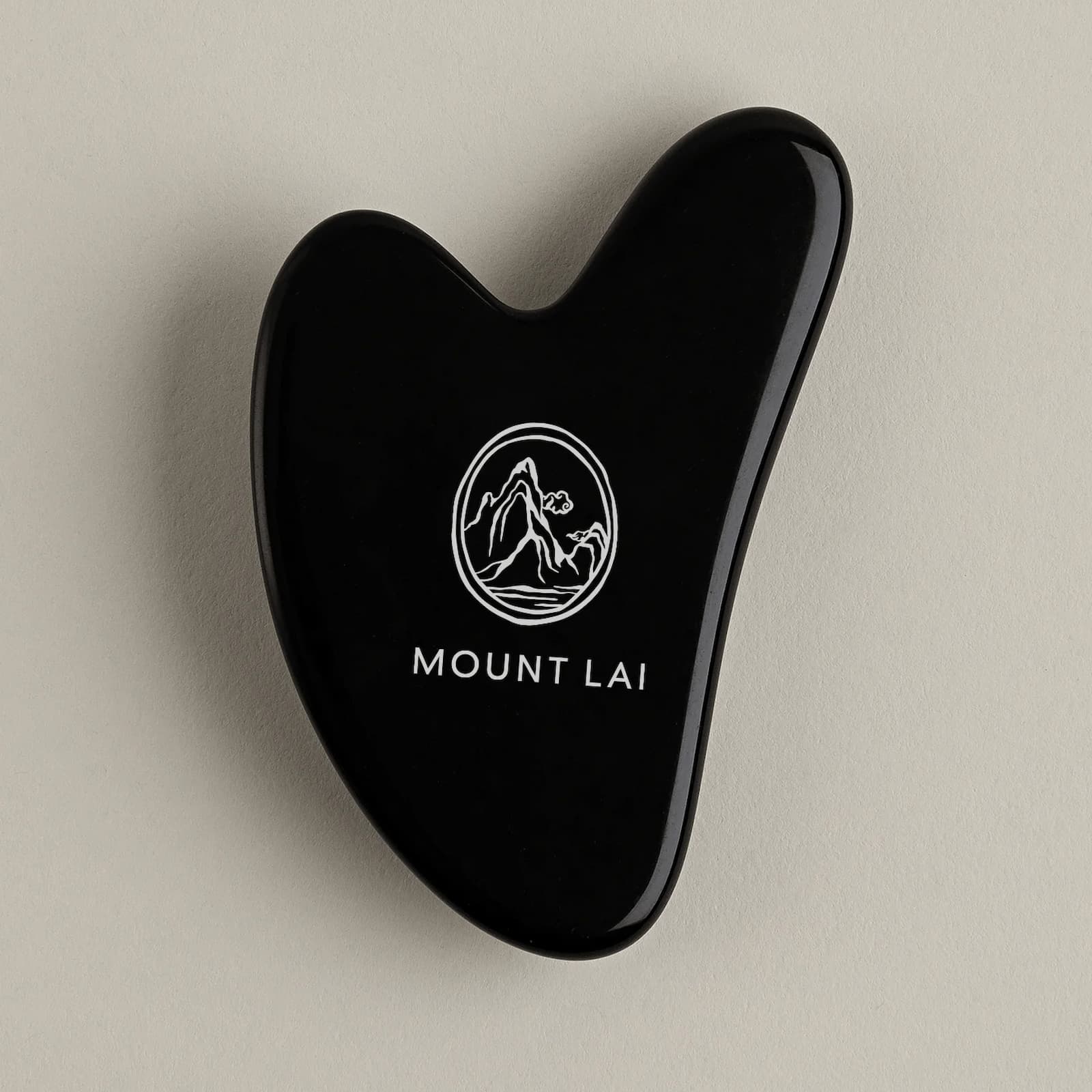 A glossy black Mount Lai Obsidian Gua Sha tool with the Mount Lai logo on the front, against a beige-grey background.