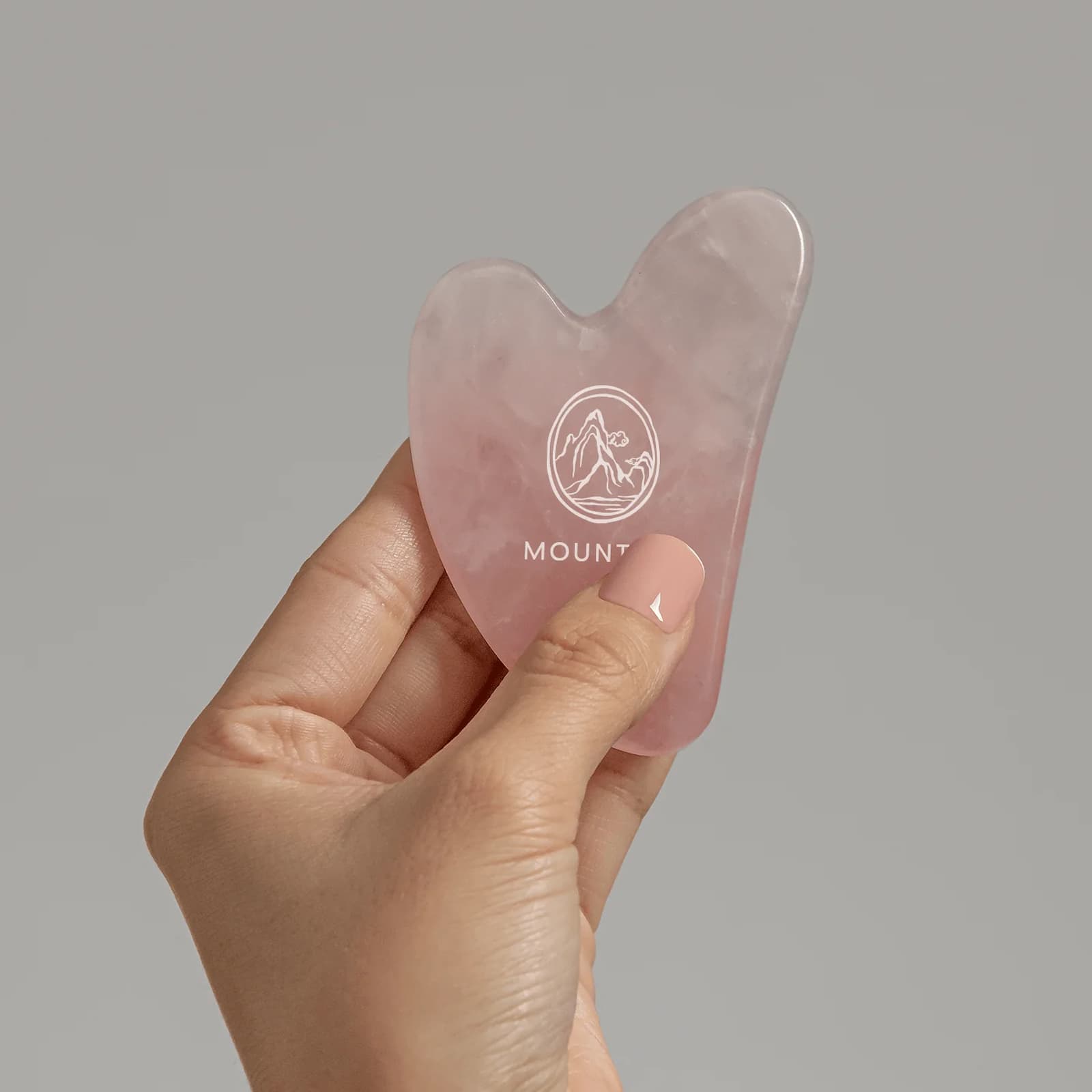 A hand holding a pink Mount Lai Rose Quartz Gua Sha tool.
