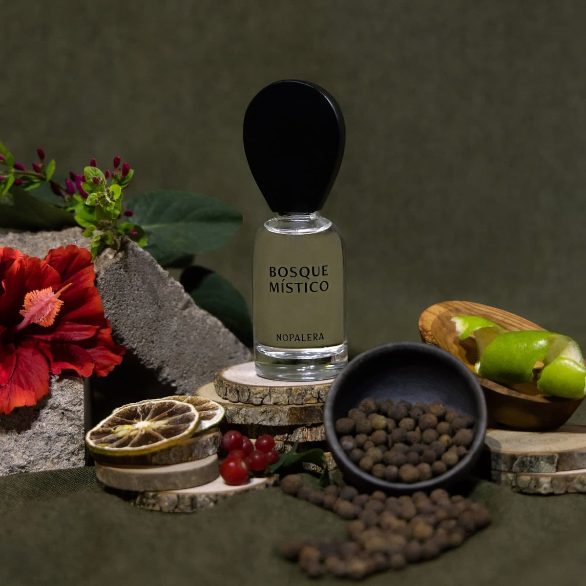A glass bottle of Nopalera "Basque Mistico" perfume sat on pieces of wood, surrounded by ingredients, against a dark green background.
