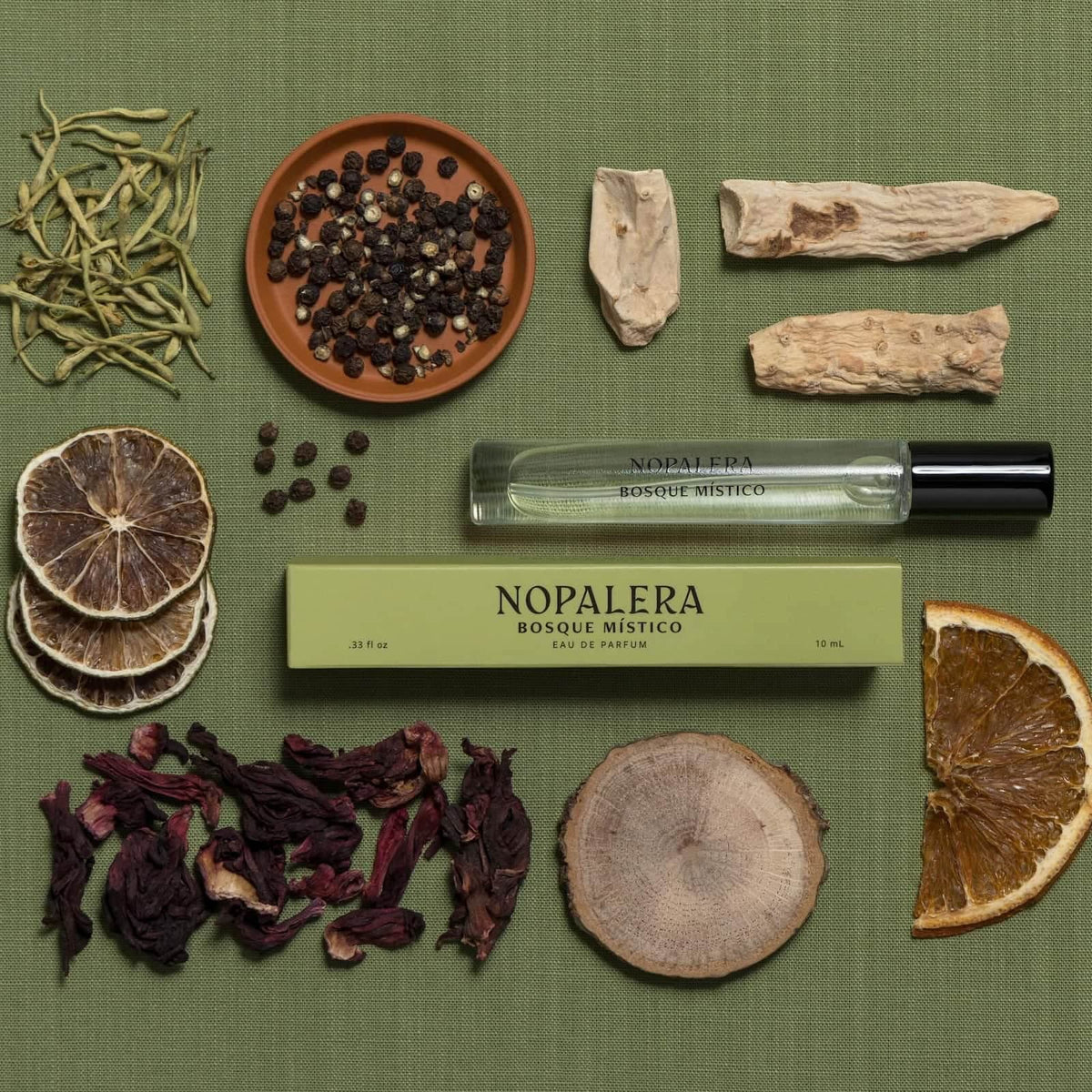 A travel-size glass bottle and green box of Nopalera "Basque Mistico" perfume against a green fabric background. Surrounded by various ingredients, including dried citrus, peppercorn, hibiscus, and wood.