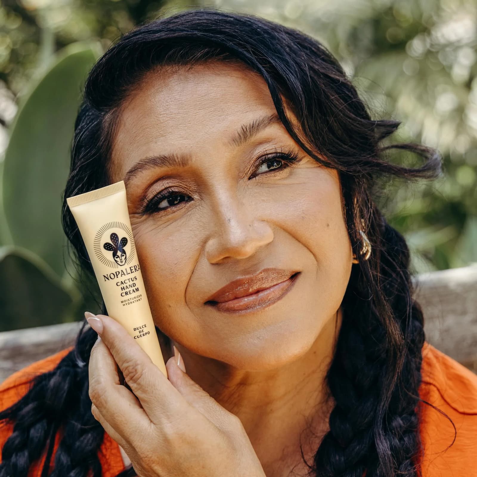 A woman in orange holding a yellow tube of Nopalera Cactus Hand Cream in Dulce de Cuerpo up to her face.