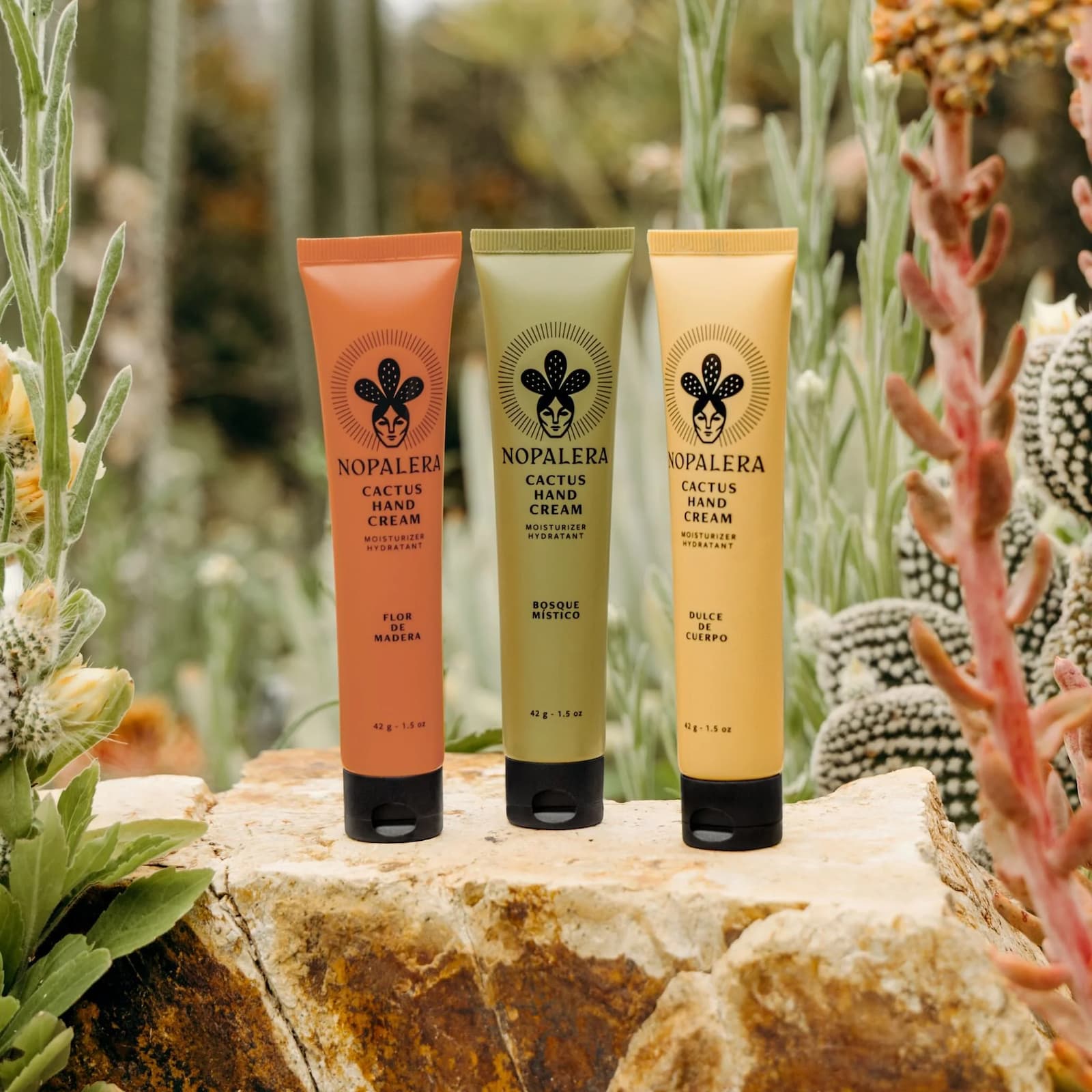 Three different colored tubes of Nopalera Cactus Hand Cream stood upright on a rock. From left to right: orange Flor de Madera, green Bosque Mistico, yellow Dulce de Cuerpo.