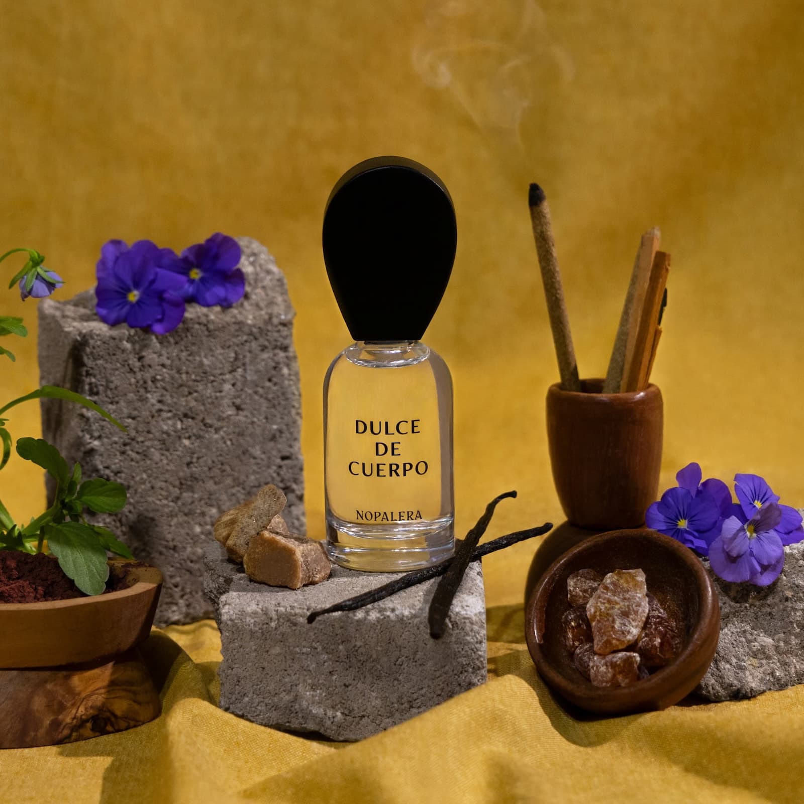 Glass bottle of Nopalera Dulce de Cuerpo sat on top of a grey stone, surrounded by various ingredients such as vanilla bean, amber, violet, and sandalwood on a yellow fabric backdrop.