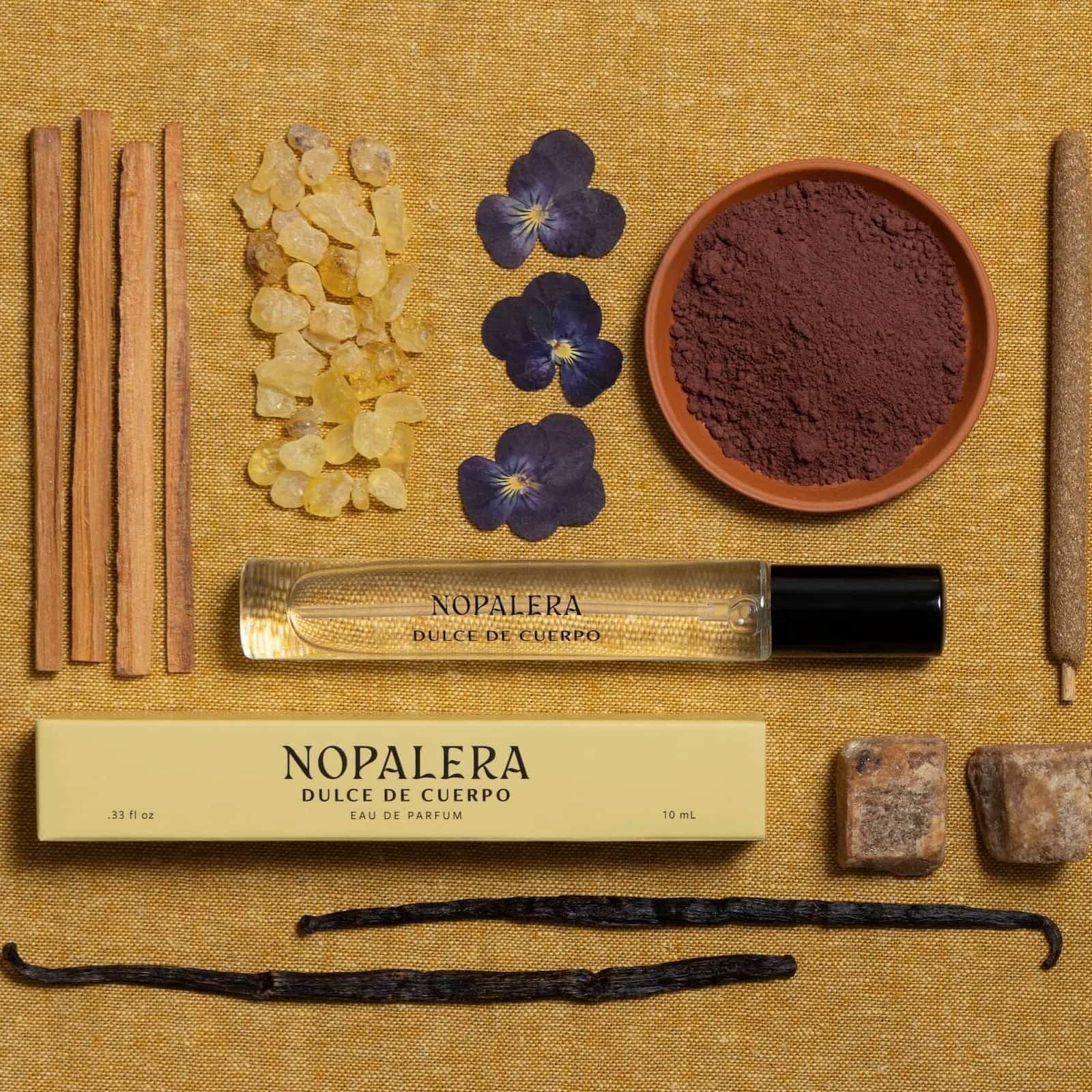 Travel-sized glass bottle of Nopalera Dulce de Cuerpo perfume laid against a yellow fabric, accompanied by the ingredients of the fragrance,
