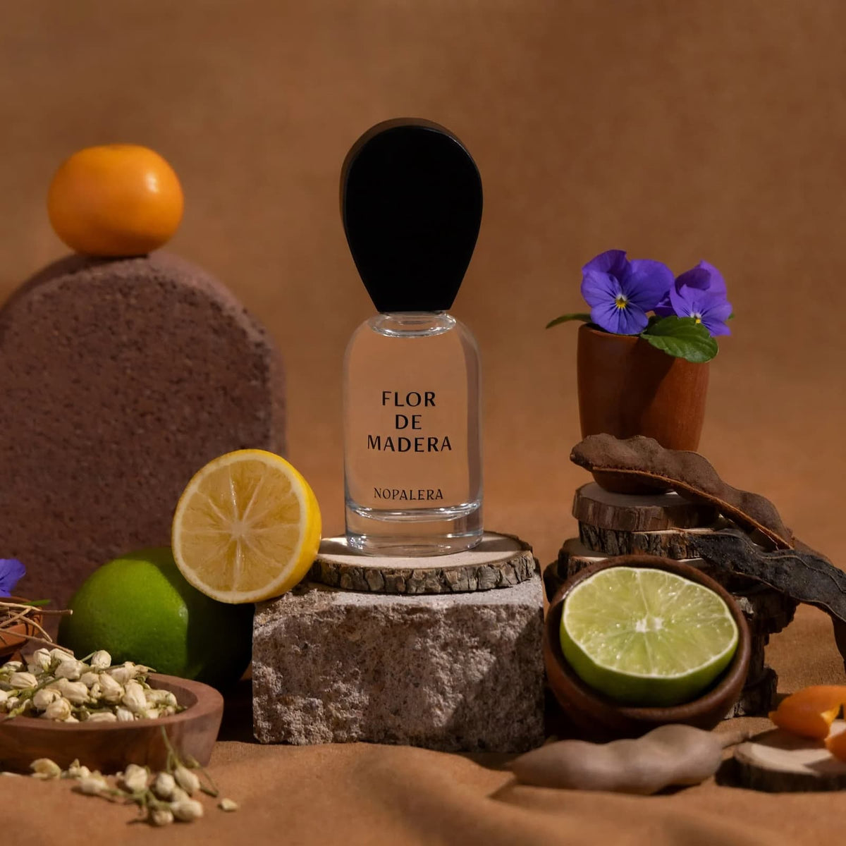 A glass bottle of Nopalera "Flor de Madera" perfume sitting on top of a piece of wood, surrounded by fragrance ingredients, including citrus, violet, and jasmine.