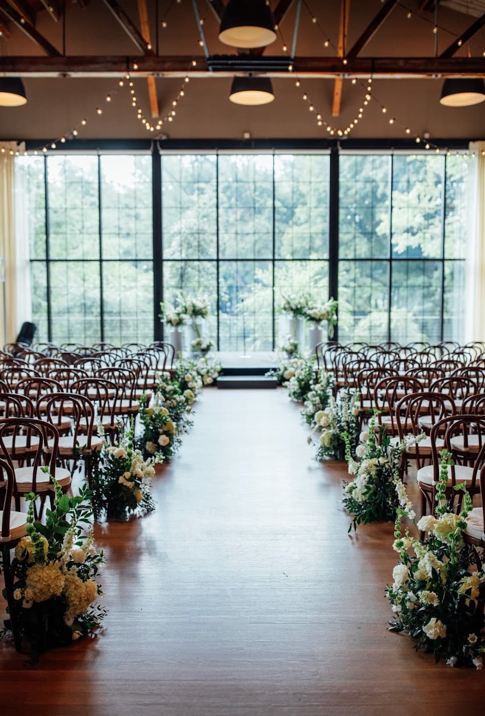A wedding ceremony setup at the Roundhouse in Beacon, NY, with rows of wooden chairs on both sides of a wooden aisle, decorated with white and green floral arrangements. Large black-framed windows in the back show green trees outside, and string lights hang from the ceiling.