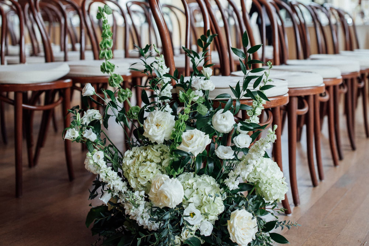 A large floral arrangement of white roses, hydrangeas, and green foliage by Witch Hazel sits on the floor between rows of wooden wedding chairs at the Roundhouse in Beacon, NY.