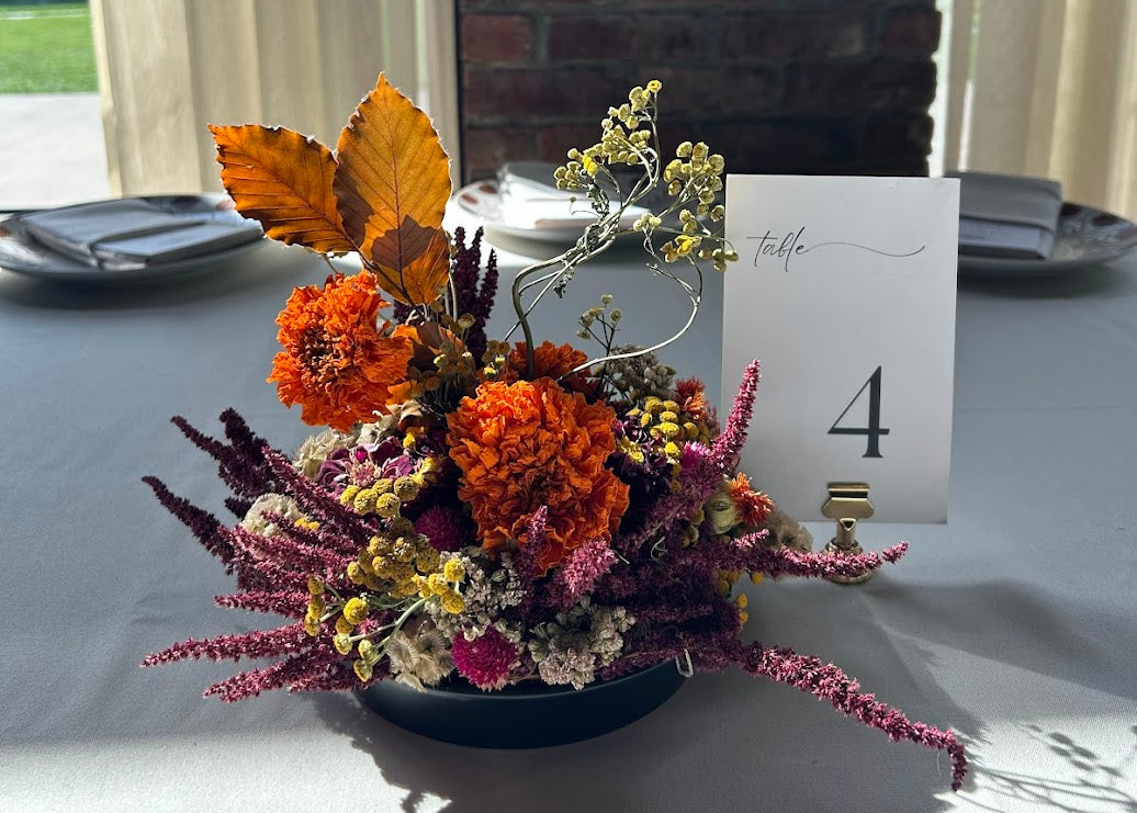 A fall Roundhouse wedding floral table centerpiece in a round black bowl, featuring orange marigolds, burgundy amaranthus, yellow tansy, and dried leaves, set on a white tablecloth next to a white table number 4.