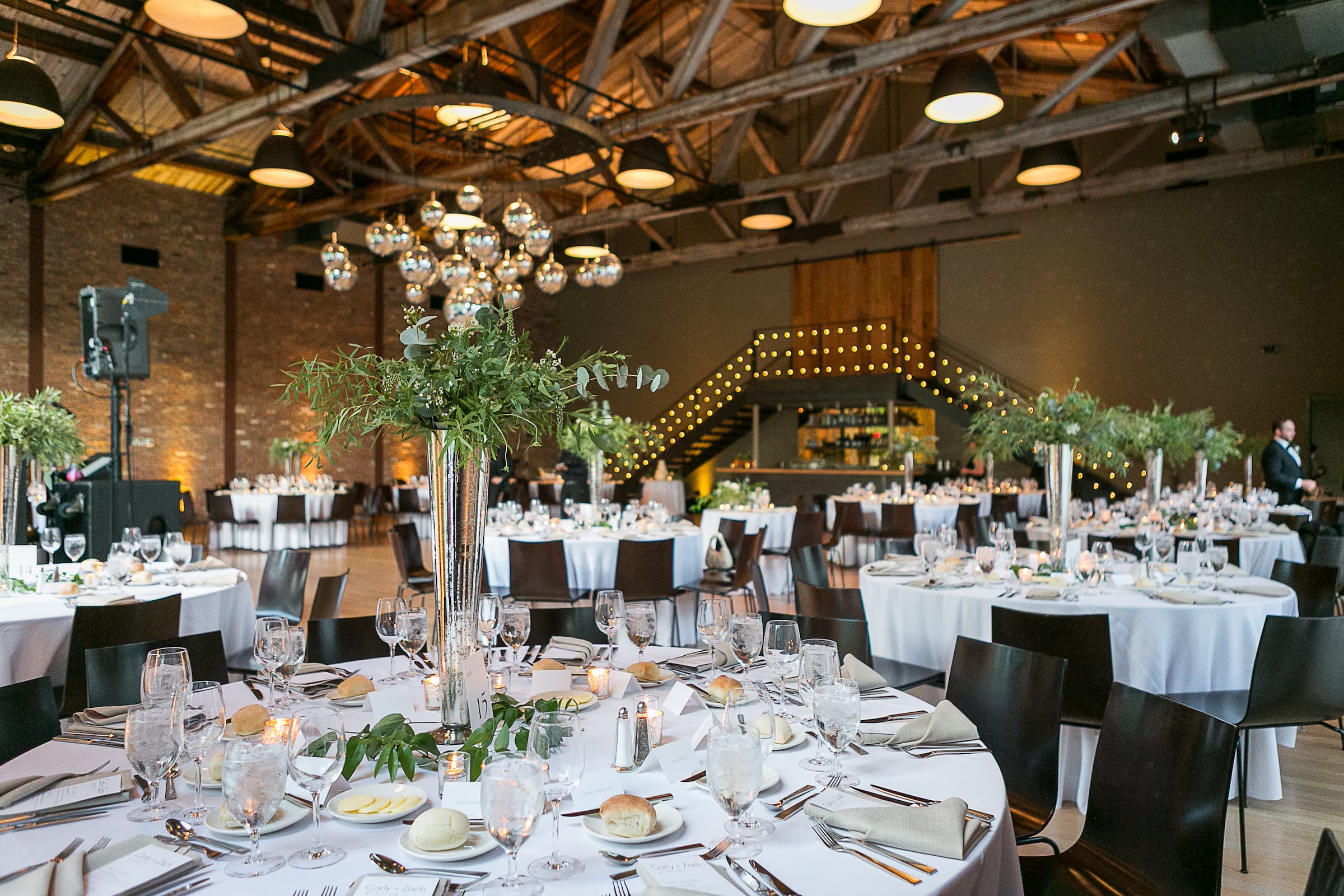 The Roundhouse reception room in Beacon, NY, with round tables set in white tablecloths, glassware, silverware, and tall greenery centerpieces. The venue has exposed wooden ceiling beams, hanging disco balls, brick walls, and warm string lights in the background.