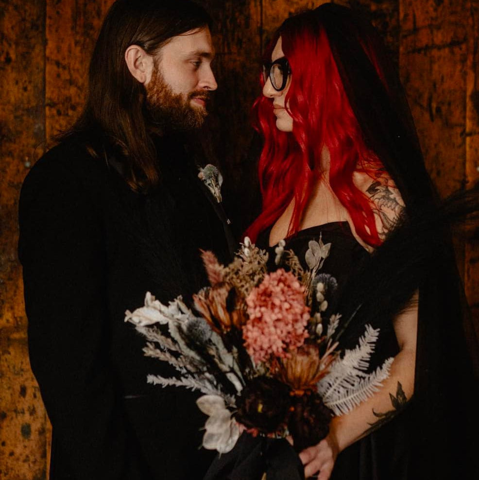 A couple in black gothic wedding attire holding a dried floral bouquet against a wooden wall. The woman has vibrant red hair and wears glasses, and the man has long dark hair and a beard.