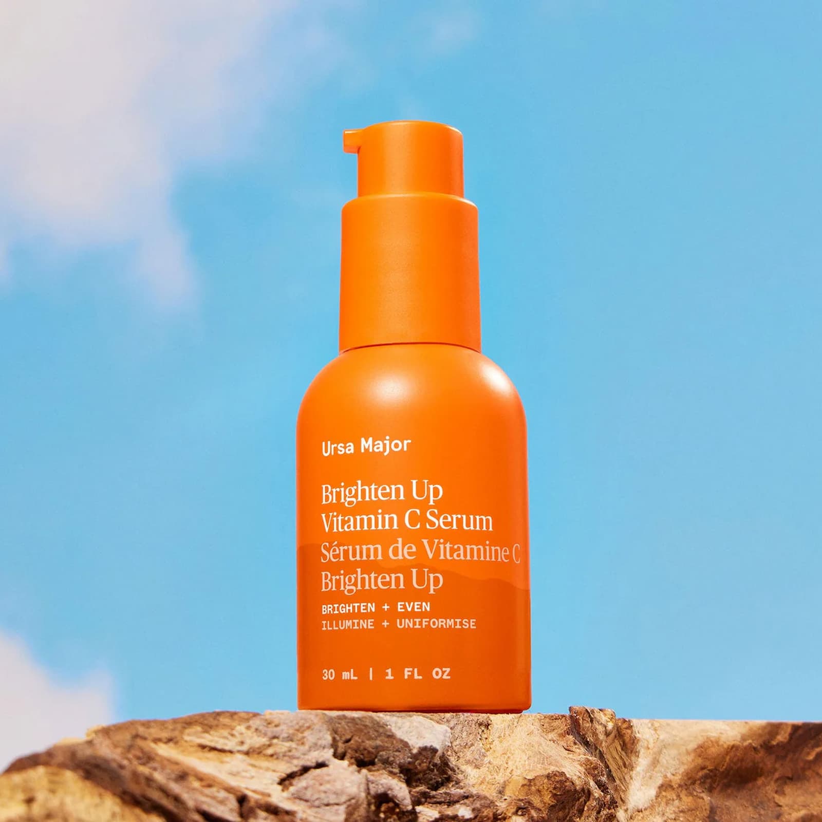 An orange bottle of Ursa Major Brighten Up Vitamin C Serum sat on a rock, in front of a blue sky background.