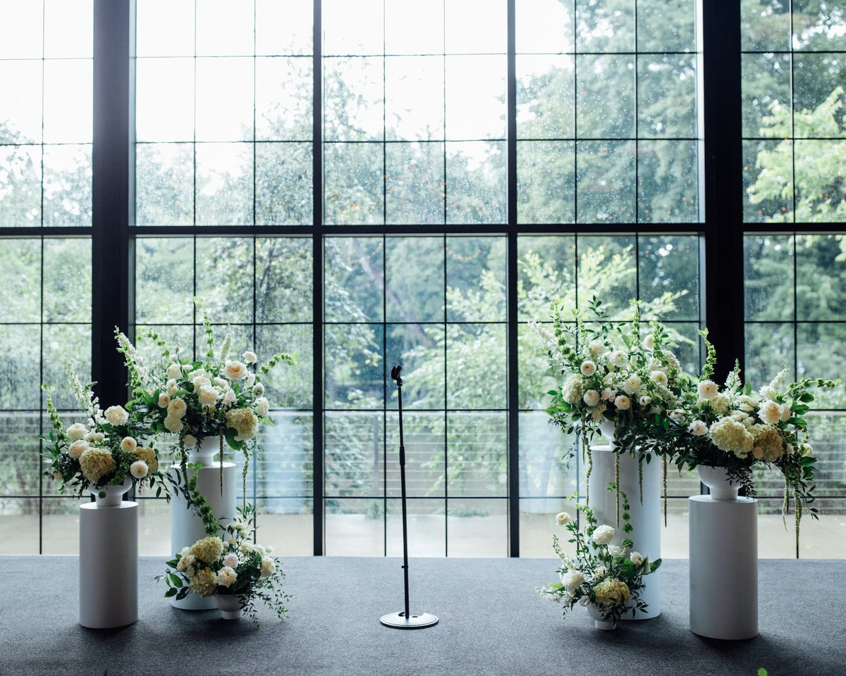 Roundhouse wedding flowers in Beacon, NY. Four white pedestals topped with white and green floral arrangements on a dark gray bandstand in front of black-framed windows overlooking green trees.